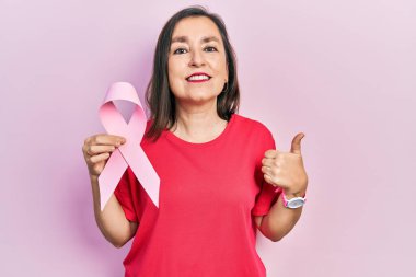 Orta yaşlı İspanyol kadın elinde pembe kanser kurdelesi, mutlu ve pozitif gülümsüyor, başparmağı mükemmel ve onay işareti yapıyor. 
