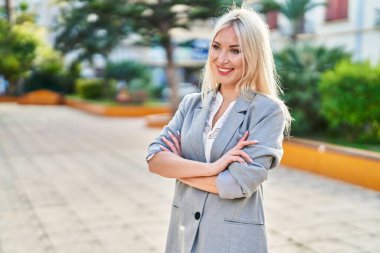 Genç sarışın kadın gülümsüyor. Parkta kollarını kavuşturmuş duruyor.