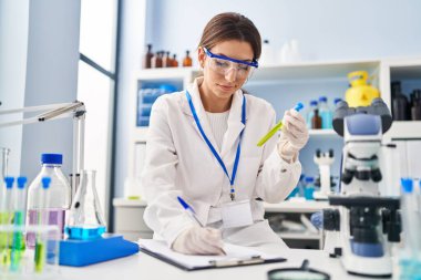 Laboratuvarda test tüpü tutan ve panoya yazı yazan bilim adamı üniforması giyen İspanyol genç bir kadın.