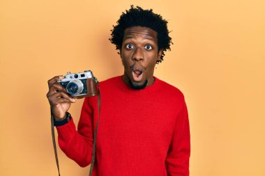 Young african american man holding vintage camera scared and amazed with open mouth for surprise, disbelief face 