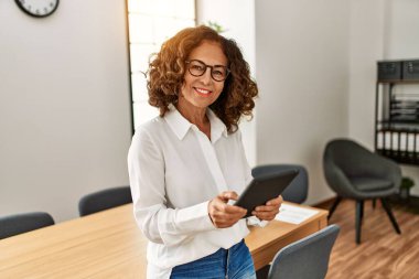 Orta yaşlı İspanyol kadın, ofiste dokunmatik ped kullanarak çalışırken kendine güvenerek gülümsüyor.
