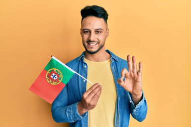 Portekiz bayrağı tutan genç Arap adam parmaklarıyla imza atıyor, gülümseyerek mükemmel bir sembol oluşturuyor. 