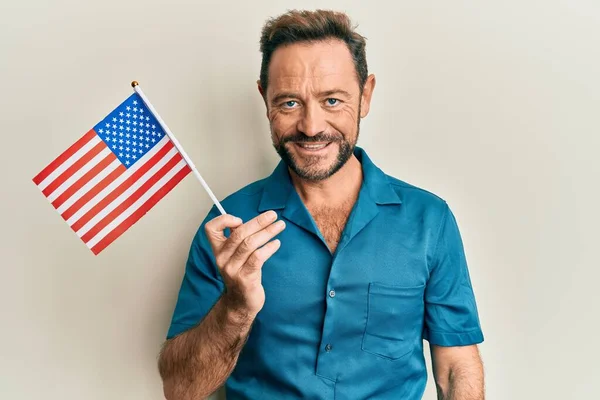 Middle age man holding united states flag looking positive and happy standing and smiling with a confident smile showing teeth