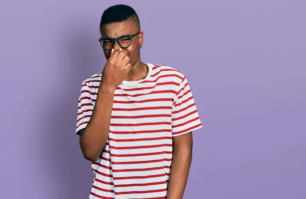 若いアフリカ系アメリカ人の男性がカジュアルなTシャツと眼鏡を着て 臭いと嫌な 耐えられない匂いを嗅ぎ 鼻の上に指で息を保持します 臭いが悪い — ストック写真