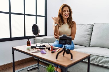 Orta yaşlı İspanyol kadın evde akıllı telefonuyla makyaj dersi kaydediyordu. Gülümsüyordu ve baş parmağıyla yüzünü gösteriyordu.. 