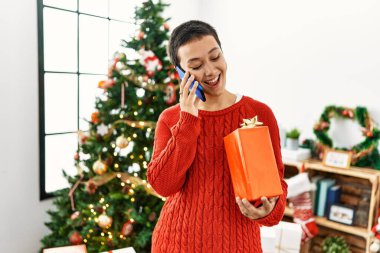 Genç İspanyol kadın elinde hediyesiyle akıllı telefondan konuşuyor. Noel ağacının yanında duruyor.