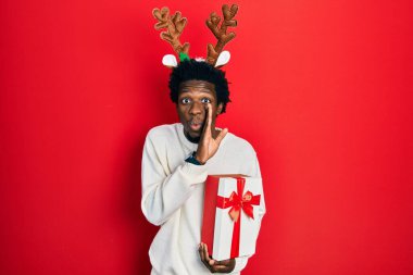 Young african american man wearing deer christmas hat holding gift hand on mouth telling secret rumor, whispering malicious talk conversation 