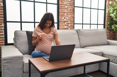 Genç Latin bir kadın hamile. Laptop kullanıyor. Evde kahve içiyor.