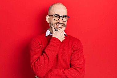 Gündelik giysiler ve gözlük takan genç kel adam kameraya kendinden emin bir şekilde bakıyor kollarını kavuşturup ve çenesini kaldırarak gülümsüyor. Olumlu düşünmek. 