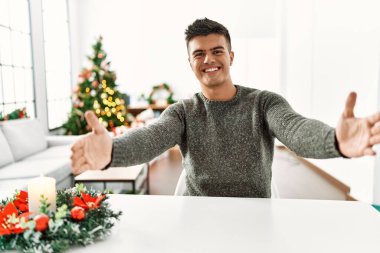 Genç İspanyol adam Noel ağacının yanında masaya oturmuş kameraya bakıyor ve sarılmak için kollarını açmış gülümsüyor. Mutluluğu kucaklayan neşeli bir ifade.. 