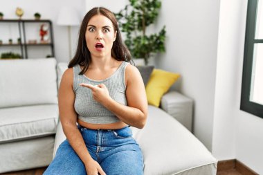 Evdeki kanepede oturan genç esmer kadın şaşkın şaşkın, ağzı açık, parmaklarını kenarı işaret ederek şaşırdı.. 