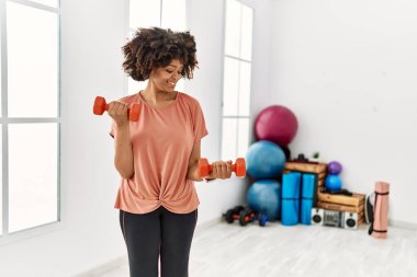 Genç Afrikalı Amerikalı kadın gülümsüyor spor merkezinde halterleri kullanarak kendine güven eğitimi alıyor