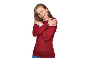 Gündelik kıyafetler giyen genç sarışın kadın mutlu ve olumlu bir şekilde kendine sarılıyor, kendinden emin gülümsüyor. Sevgi ve özgüven. 