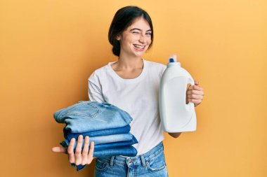 Çamaşır deterjanı şişesi ve katlanmış kot pantolon tutan genç beyaz kadın kameraya seksi bir ifadeyle bakıyor, neşeli ve mutlu bir yüzle.. 