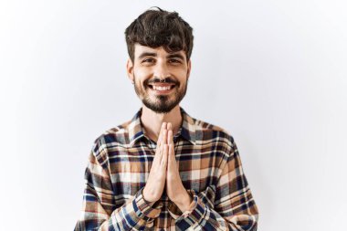 Sakallı İspanyol bir adam soyutlanmış arka planda duruyor ellerinin birarada dua ederek af diliyor kendinden emin gülümsüyor. 