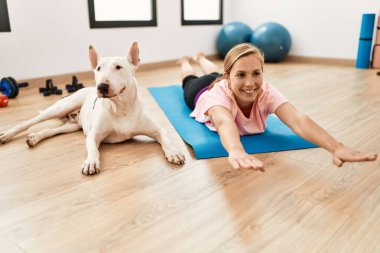 Genç beyaz kadın gülümsüyor spor merkezinde köpekle esniyor.