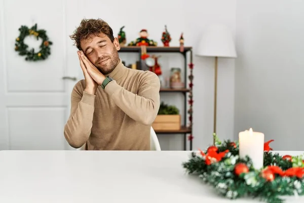 年轻英俊的男人 留着胡子 坐在桌子上 挂着圣诞装饰品 睡懒觉 手牵着手 闭着眼睛笑着 — 图库照片