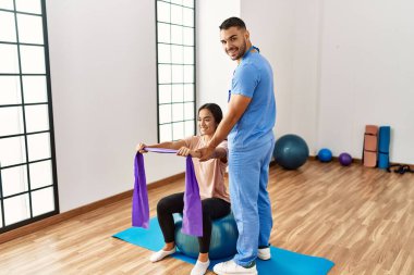 Latin erkek ve kadın fizyoterapist üniforması giyiyorlar rehabilitasyon merkezinde uygun top ve elastik bant kullanarak rehabilitasyon seansı yapıyorlar.