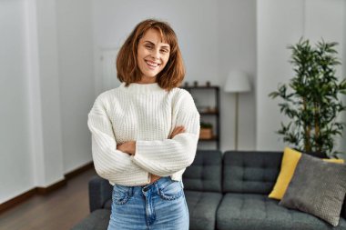 Beyaz bir kadın gülümsüyor. Kollarını kavuşturup evde duruyor.