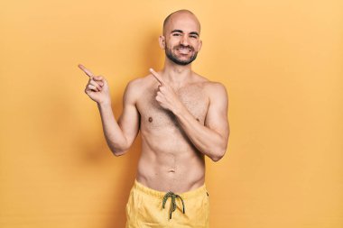 Mayolu genç kel adam gülümsüyor ve kameraya bakıyor. İki eliyle ve parmaklarıyla kenarı işaret ediyor.. 