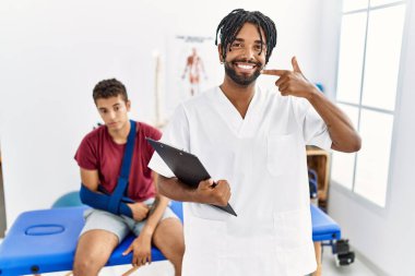 Kırık kollu, neşeli gülümseyen ve parmaklarıyla ve ağzıyla işaret eden bir adamla birlikte ağrı giderme kliniğinde çalışan genç İspanyol bir adam. diş sağlığı kavramı. 