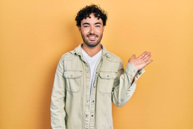 Gündelik kıyafetler giyen genç İspanyol adam neşeli bir şekilde gülümsüyor ve avuç içi kameraya bakıyor.. 