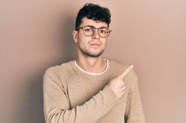 Gündelik kıyafetler giyen ve gözlüklü genç İspanyol adam el parmağı ile yan tarafı işaret ediyor. Ciddi ve sakin bir yüz ifadesi sergiliyor. 