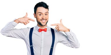 Sakallı, hippi görünümlü, papyonlu, pantolon askısı takan neşeli gülümsemeli ve parmakları, dişleri ve ağzıyla işaret eden İspanyol bir adam. diş sağlığı kavramı. 