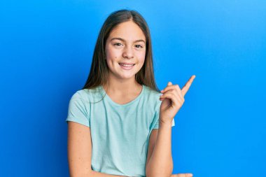 Güzel, esmer, beyaz tişörtlü, yüzünde kocaman bir gülümseme olan küçük bir kız. Eli ve parmağı kameraya bakıyor.. 