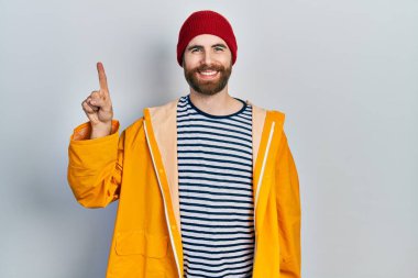 Sakallı, sarı yağmurluk giyen beyaz bir adam bir numaralı parmağını gösterip işaret ederken kendinden emin ve mutlu bir şekilde gülümsüyor.. 