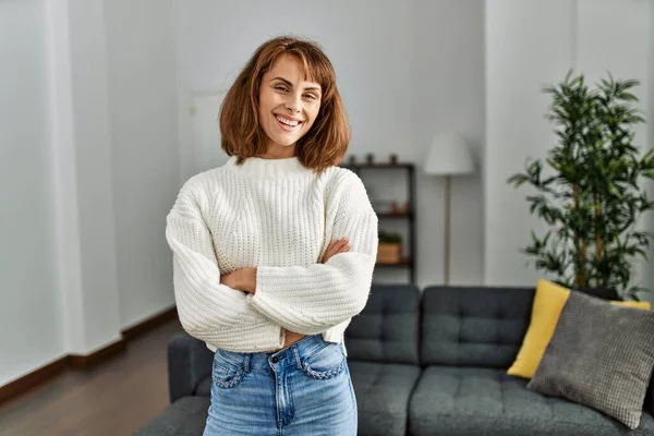 Beyaz bir kadın gülümsüyor. Kollarını kavuşturup evde duruyor.