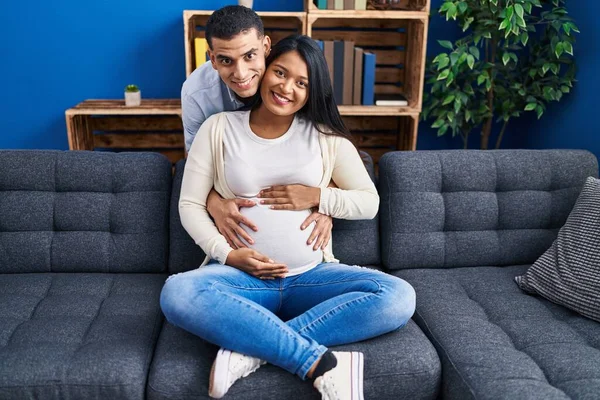 Young Latin Couple Expecting Baby Hugging Each Other Sitting Sofa — Stock fotografie