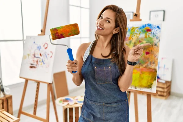 若いですラテンアーティスト女性保持ペイントローラースタンドでアートスタジオ笑顔幸せなポインティングとともに手と指で側面 — ストック写真