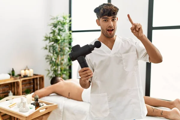 Young Hispanic Man Holding Therapy Massage Gun Wellness Center Surprised — Stock Photo, Image
