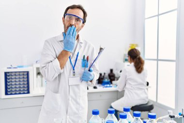 Orta yaşlı bir adam laboratuvarda çalışıyor. Ağzını eliyle kapatıyor. Şok geçiriyor ve hata yapmaktan korkuyor. şaşırmış ifade 