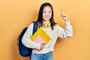 Sırt çantası ve kitapları tutan Çinli genç bir kız. Şaşırmış, şaşırmış, parmaklarıyla işaret ediyor ve kollarını kaldırıyordu.. 