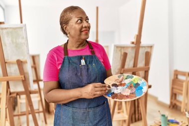Kıdemli Afro-Amerikan kadın sanat stüdyosunda palet ve boya fırçası tutarak gülümsüyor.
