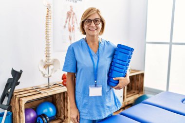 Klinikte köpük silindir tutan fizyoterapist üniforması giyen orta yaşlı sarışın bir kadın.