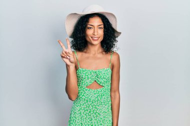Genç Latin kız yaz şapkası takıyor ve iki numarayı gösterip işaret ederken kendinden emin ve mutlu gülümsüyor. 