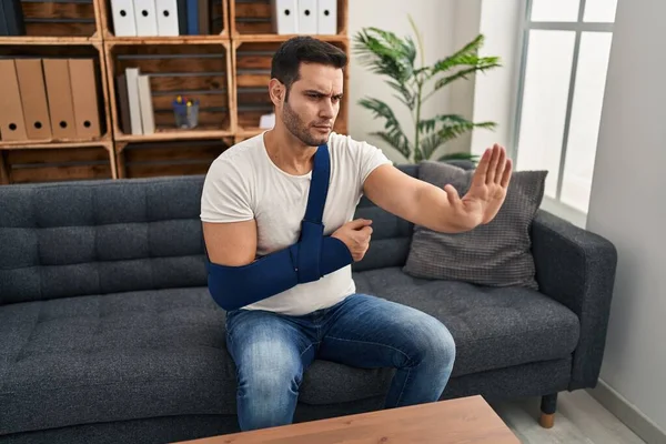 Young hispanic man with beard wearing arm on sling sitting at therapy consult with open hand doing stop sign with serious and confident expression, defense gesture