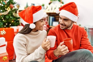 Mutlu gülümseyen ve kahve içen genç İspanyol çift evdeki Noel ağacının yanında yerde oturuyor..