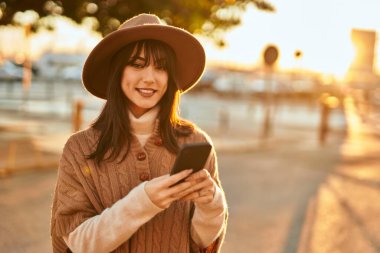 Kışlık şapkalı esmer kadın gün batımında şehrin dışında akıllı telefon kullanarak gülümsüyor.