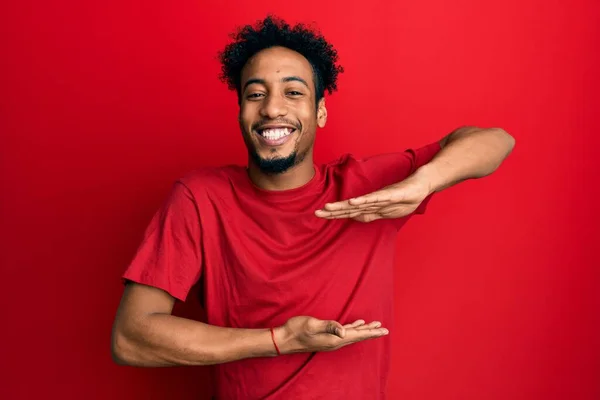 年轻的非洲裔美国人 留着胡子 身穿宽松的红色T恤 手拿着大号和大号的手势 象征着什么 微笑着看着摄像机 计量概念 — 图库照片