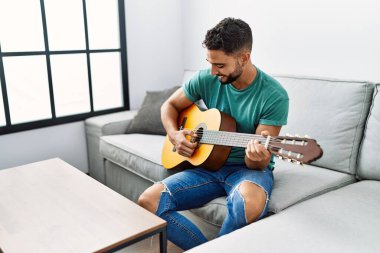 Genç Arap adam evdeki kanepede oturup klasik gitar çalıyor.