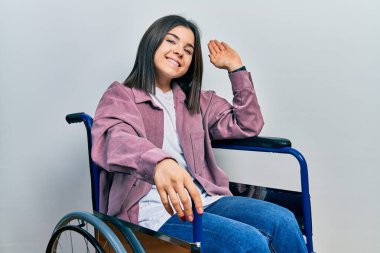 Tekerlekli sandalyede oturan esmer kadın selam veriyor mutlu ve gülümsüyor, dostça karşılama hareketi yapıyor. 