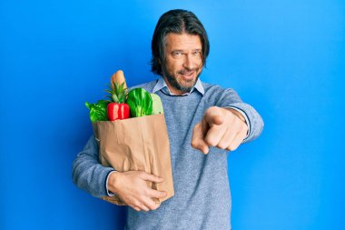 Orta yaşlı, elinde alışveriş poşeti olan yakışıklı bir adam kamerayı işaret ediyor. Kendine güvenen bir jest, ciddi görünüyor. 