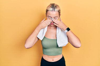 Spor kıyafetleri ve havluyla gezen genç sarışın kız yorgunluk ve baş ağrısı için gözlerini ovalıyor, uykulu ve yorgun bir ifade takınıyor. görme sorunu 