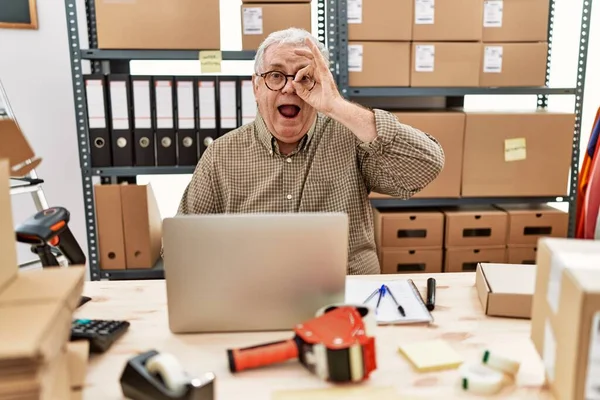 Senior caucasian man working at small business ecommerce with laptop doing ok gesture shocked with surprised face, eye looking through fingers. unbelieving expression.