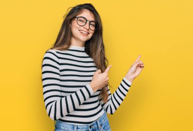 Genç ve güzel bir kız, günlük kıyafetler ve gözlük takıyor gülümsüyor ve kameraya bakıyor iki eliyle ve parmaklarıyla kenarı işaret ediyor.. 