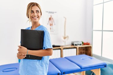 Genç beyaz fizyoterapist klinikte mutlu bir şekilde klasörü tutuyor.